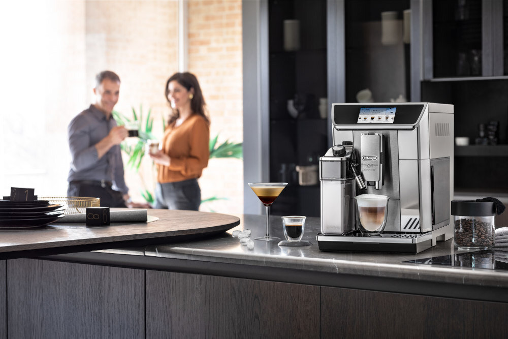 Na blacie kuchennym stoi srebrny ekspres do kawy z logo \'DeLonghi\', obok którego znajdują się filiżanki z kawą. W tle widać dwie osoby rozmawiające przy oknie.