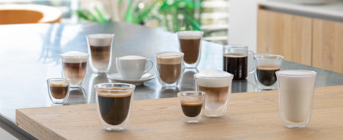 Na drewnianym blacie stoi kilka filiżanek i szklanek z różnymi rodzajami kawy, w tym espresso, cappuccino i latte. W tle widoczne są zielone rośliny i fragmenty nowoczesnej kuchni.