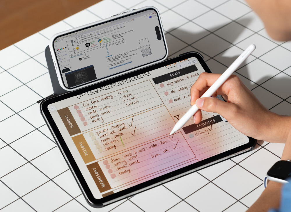 Na stole z białą kratką leży tablet z otwartą aplikacją do planowania, obok którego stoi telefon w etui. Osoba trzyma rysik i pisze na ekranie tabletu.