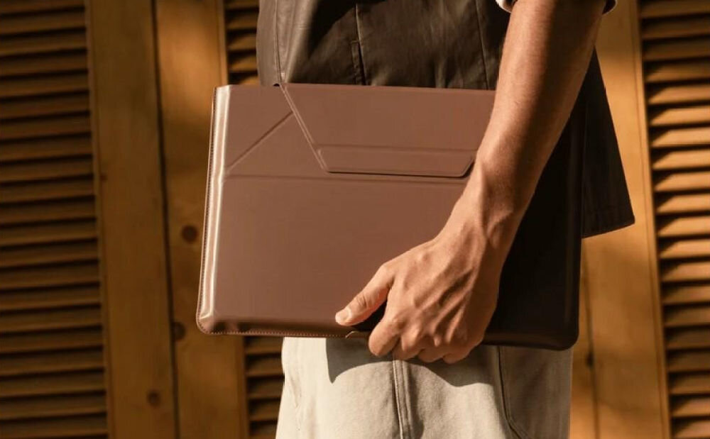 Osoba trzyma ciemnobrązową torbę na laptopa, stojąc przed drewnianą ścianą z żaluzjami. Torba ma prosty, elegancki design.
