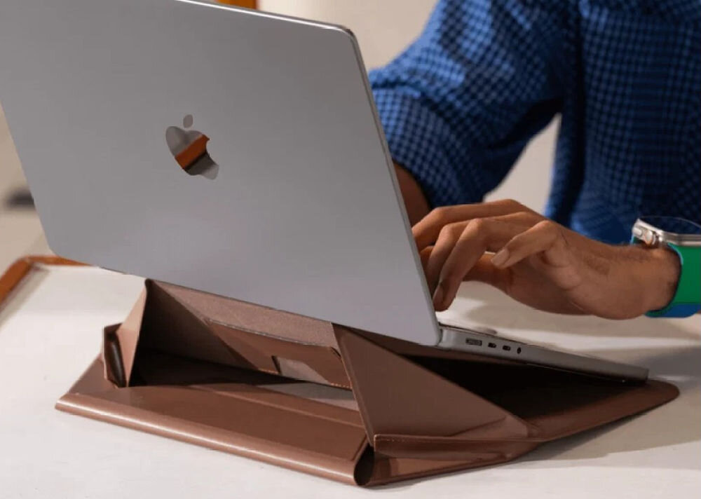 Osoba używa laptopa umieszczonego na ciemnobrązowej podstawce, która pełni funkcję torby na laptopa. Na laptopie widoczne jest logo w kształcie nadgryzionego jabłka.