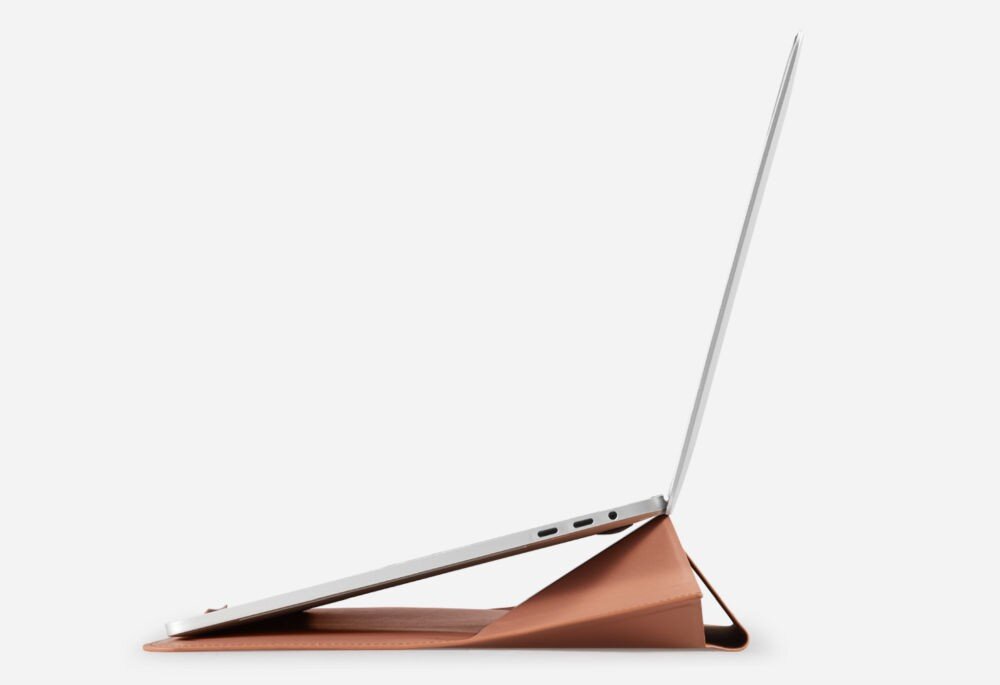 Na obrazie widoczny jest laptop ustawiony na brązowej podstawce, która unosi jego tył. Tło jest jednolite i jasne.