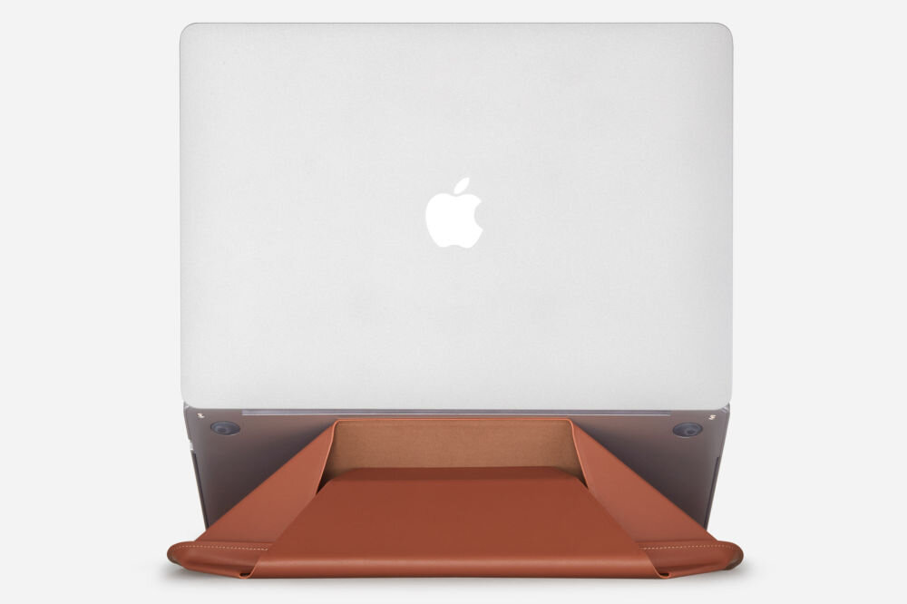 Na obrazie widoczny jest laptop z logo w kształcie nadgryzionego jabłka, ustawiony na podstawce w kolorze ciemnego brązu. Podstawka ma kształt trójkątny i jest złożona w sposób umożliwiający podparcie laptopa.