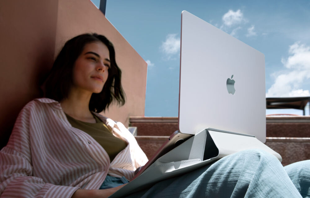 Osoba siedzi na schodach z laptopem na kolanach, na którym widoczne jest logo w kształcie nadgryzionego jabłka. Laptop jest podparty szarym stojakiem, a w tle widać fragment budynku i niebo.
