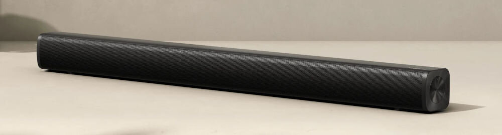 Czarny soundbar o prostokątnym kształcie z delikatnie zaokrąglonymi krawędziami, umieszczony na jasnym tle. Powierzchnia urządzenia ma teksturowane wykończenie.