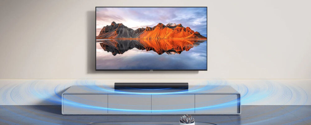 Na obrazie widać telewizor z wyświetlonym krajobrazem górskim oraz czarny soundbar umieszczony na szafce pod telewizorem. Wokół soundbara widoczne są niebieskie linie symbolizujące dźwięk.