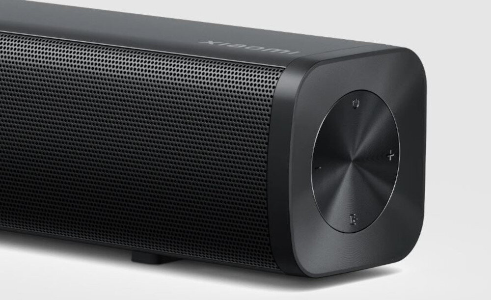 Czarny soundbar z metaliczną siatką na froncie i okrągłym panelem sterowania z przyciskami plus i minus. Na górze widoczne jest logo \'xiaomi\'.