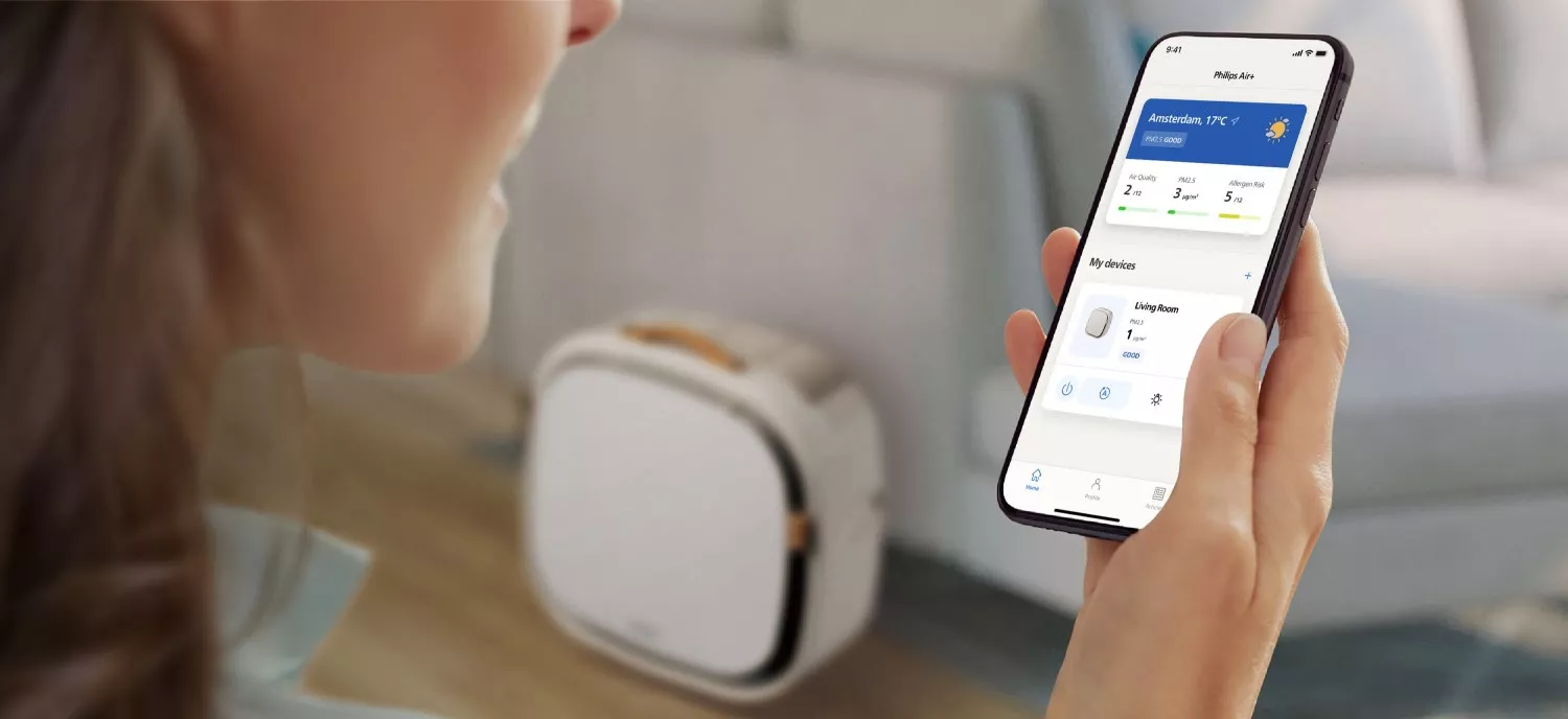 Osoba trzyma smartfon z aplikacją pokazującą informacje o jakości powietrza i urządzeniach. W tle widoczny jest oczyszczacz powietrza.