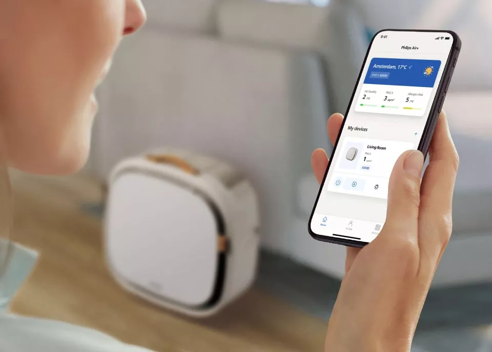 Osoba trzyma smartfon z aplikacją Philips Air, pokazującą informacje o jakości powietrza i urządzeniach. W tle widoczny jest biały oczyszczacz powietrza.