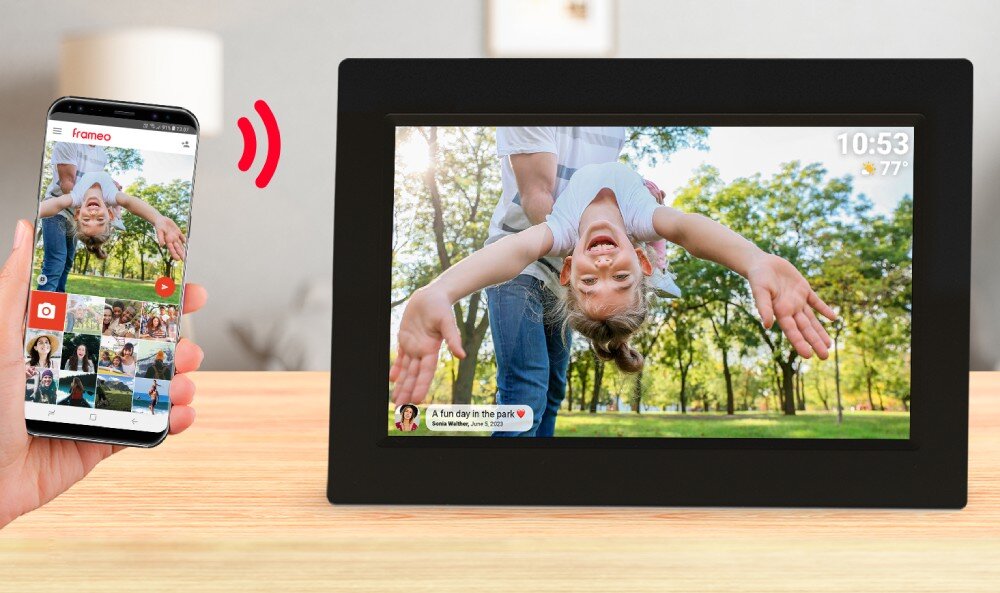 Na drewnianym blacie stoi czarna ramka cyfrowa wyświetlająca zdjęcie dziecka w parku. Obok znajduje się smartfon z aplikacją Frameo, pokazujący to samo zdjęcie i kilka miniatur innych zdjęć.