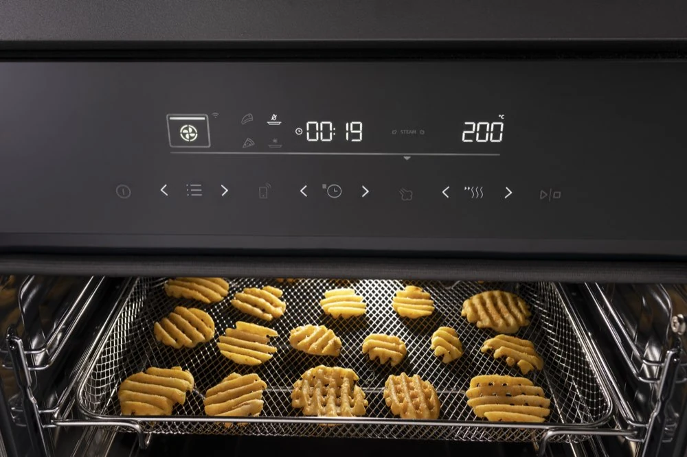 Piekarnik z wyświetlaczem pokazującym czas 00:19 i temperaturę 200°C, z widocznymi ciastkami na metalowej kratce wewnątrz. Panel sterowania zawiera różne ikony i przyciski dotykowe.