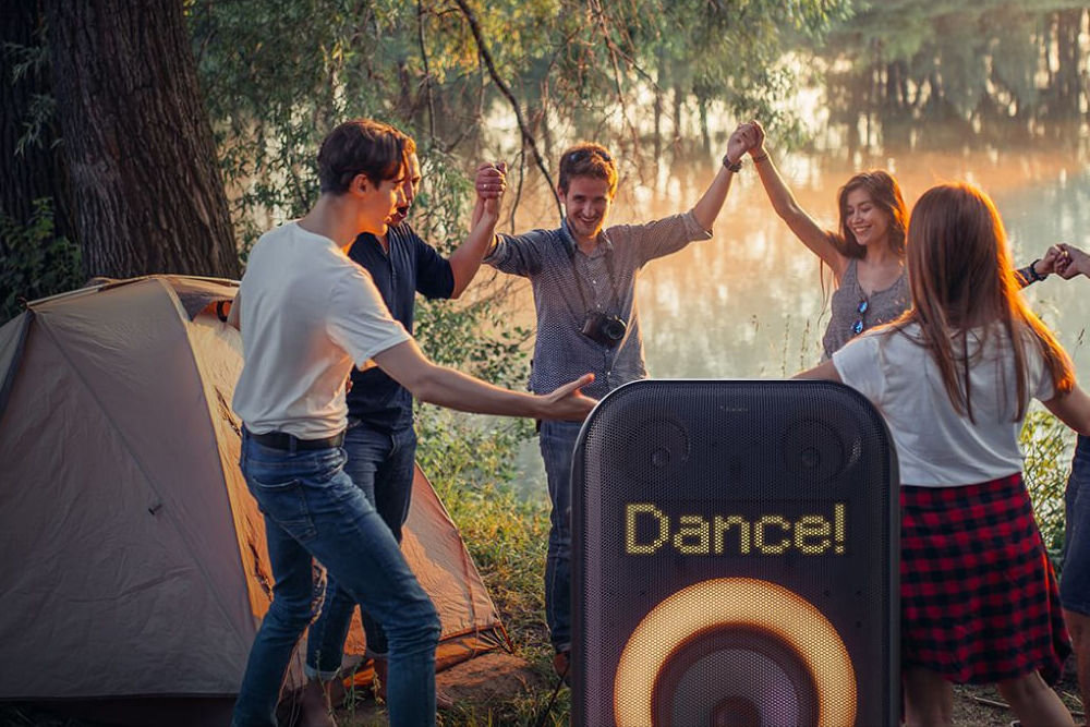 Grupa osób trzyma się za ręce obok namiotu, a na pierwszym planie widoczny jest głośnik z wyświetlaczem pokazującym napis \'Dance!\'. W tle znajduje się zalesiony teren nad wodą.