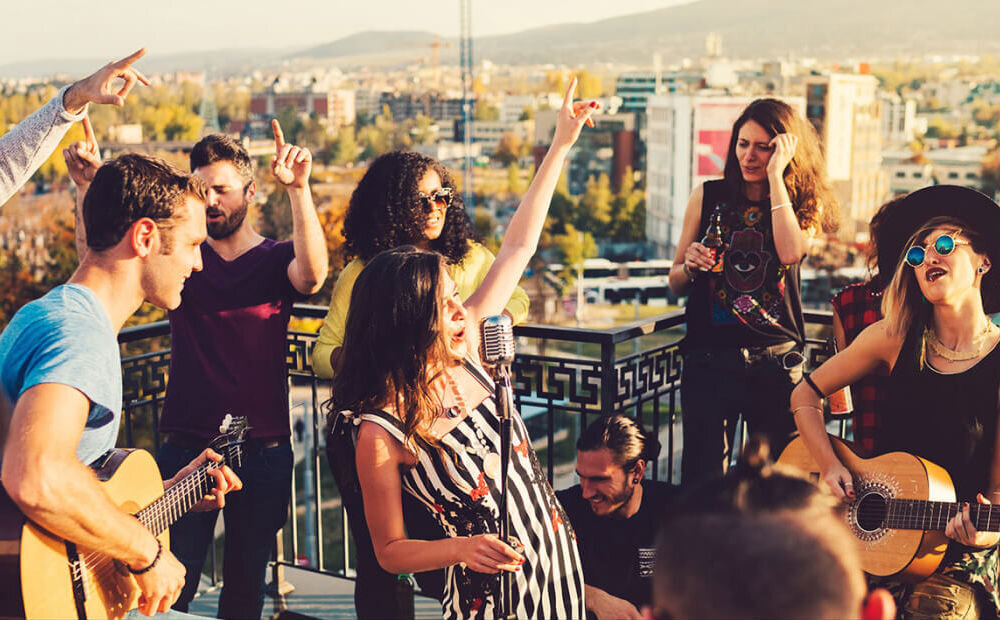 Grupa ludzi bawi się na dachu budynku, niektórzy grają na gitarach, a inni tańczą. W tle widać panoramę miasta z budynkami i wzgórzami.
