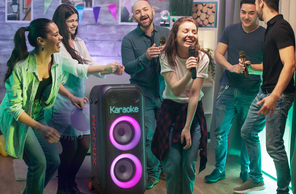 Grupa osób bawi się z mikrofonami przy dużym głośniku z napisem \'Karaoke\' i \'XBOOM\', który ma podświetlane głośniki. W tle widać dekoracje i kolorowe światła.