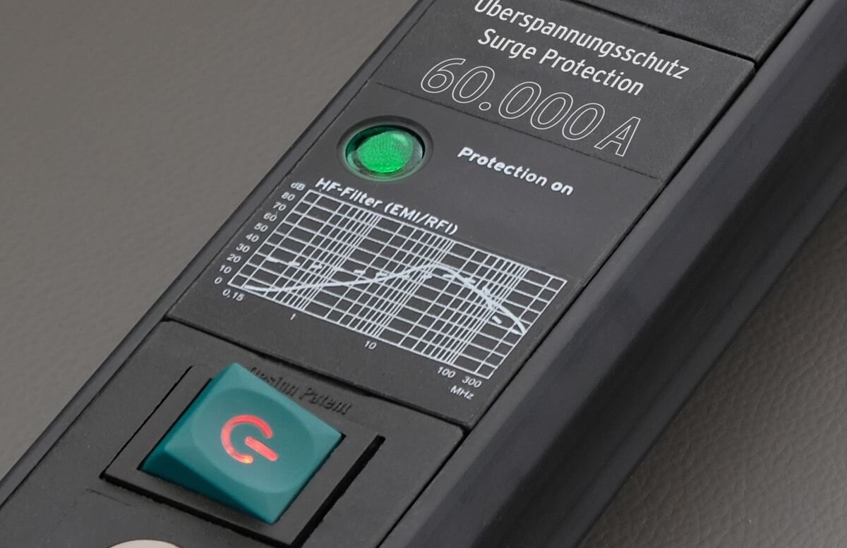 Na obrazie widoczna jest listwa zasilająca z napisem \'Überspannungsschutz Surge Protection 60.000A\'. Obok znajduje się zielona dioda z napisem \'Protection on\' oraz wykres oznaczony jako \'HF-Filter (EMI/RFI)\'.