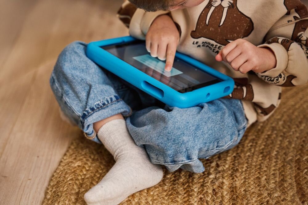 Dziecko siedzi na dywanie, trzymając niebieski tablet w ochronnym etui. Na ekranie tabletu widoczny jest tekst, który dziecko dotyka palcem.
