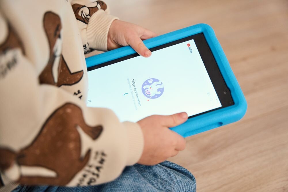 Dziecko trzyma niebieski tablet z wyświetlonym ekranem startowym aplikacji YouTube Kids. Na ekranie widoczna jest ikona z uśmiechniętą postacią i napis \'Pobieranie aplikacji\'.