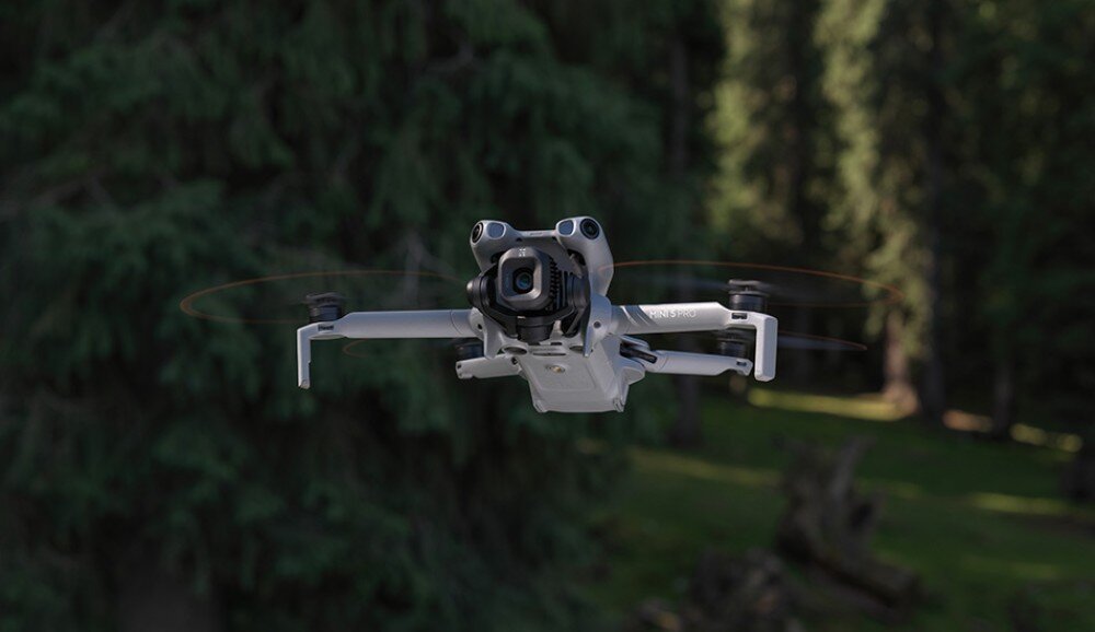 Dron w locie na tle zielonego lasu, z widocznymi czterema wirnikami i kamerą z przodu.