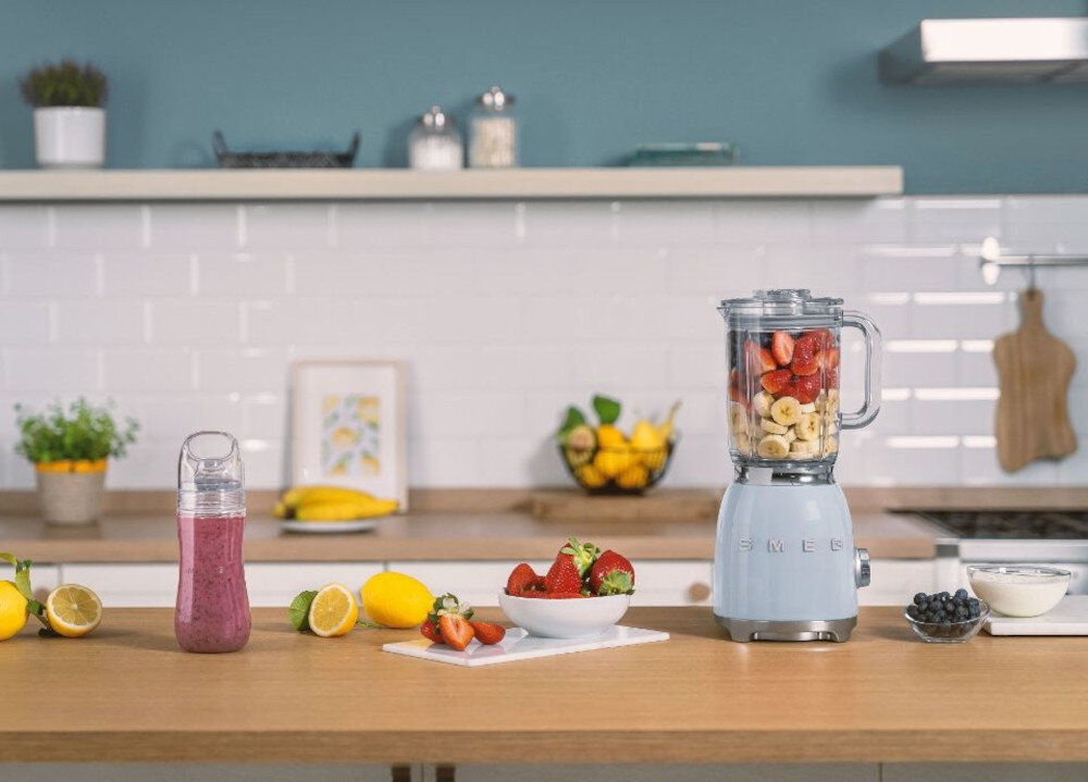 Na blacie kuchennym znajduje się blender z logo SMEG, wypełniony kawałkami owoców. Obok stoją miski z owocami, butelka z różowym napojem oraz różne owoce i jogurt.
