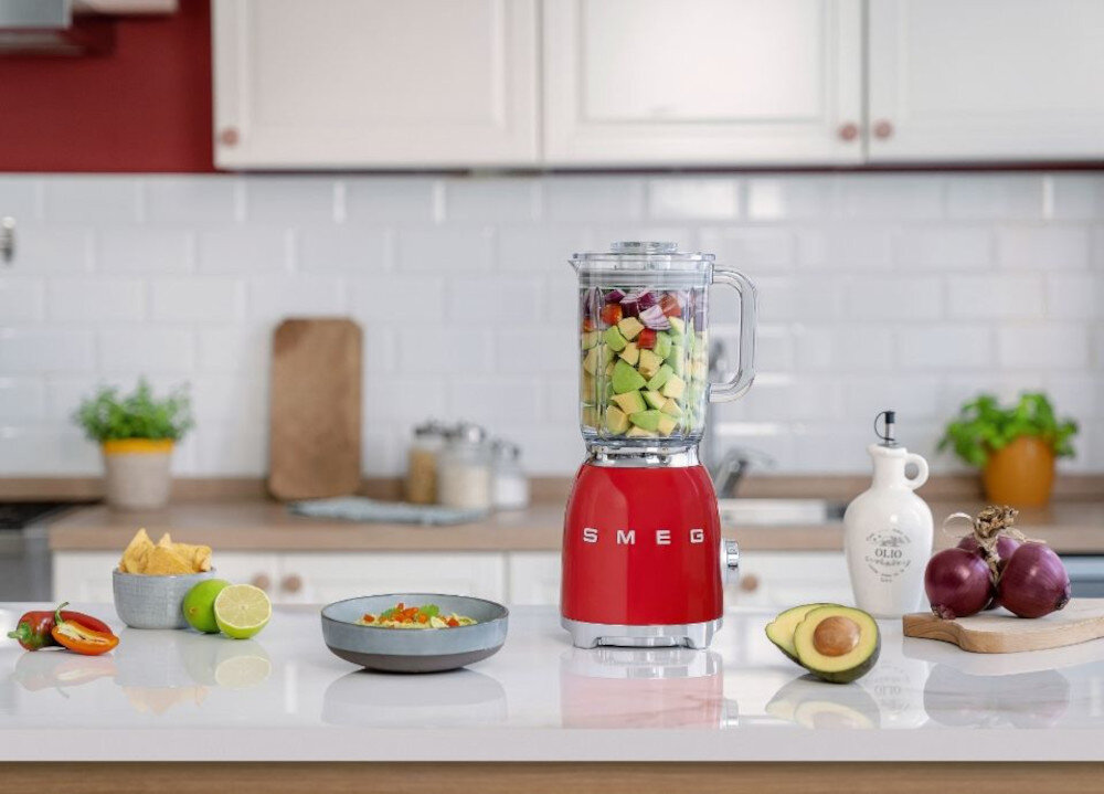 Na blacie kuchennym stoi czerwony blender z logo SMEG, wypełniony pokrojonymi owocami i warzywami. Obok znajdują się różne składniki, takie jak awokado, cebula, limonka i miska z dipem.