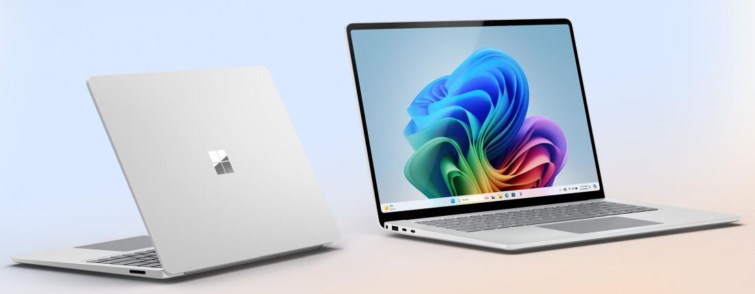 Na obrazie widoczne są dwa laptopy w srebrnym kolorze, jeden zamknięty z logo w kształcie okna, drugi otwarty z kolorowym ekranem przedstawiającym abstrakcyjny wzór. Laptopy mają cienkie ramki ekranu i minimalistyczny design.