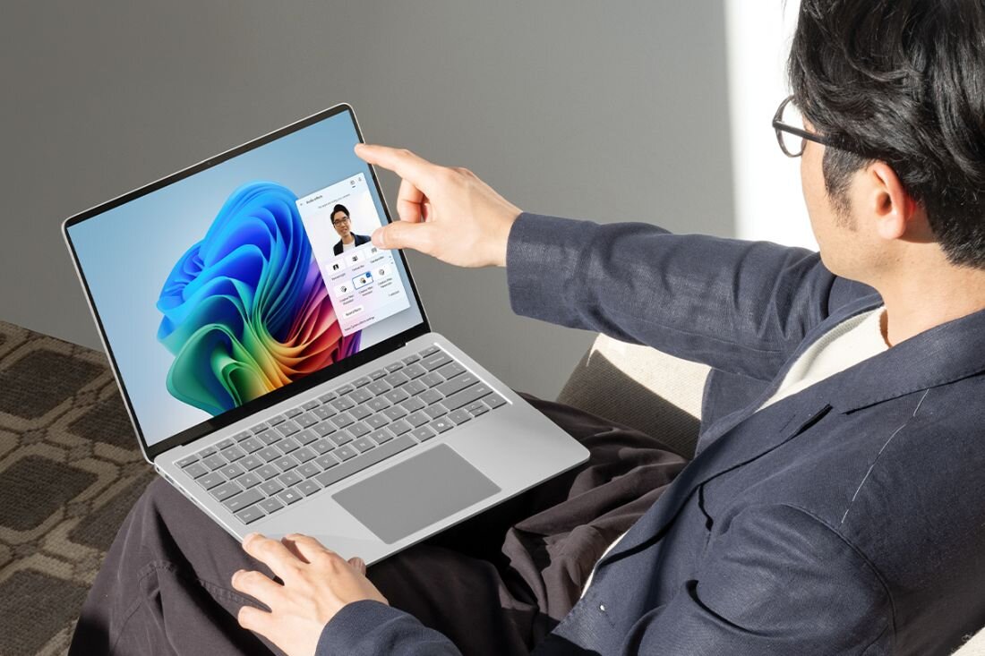 Osoba siedzi na kanapie i używa laptopa z kolorowym ekranem, na którym widoczny jest interfejs użytkownika. Laptop ma srebrną obudowę i klawiaturę.