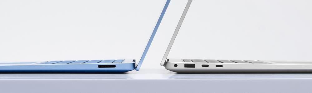 Dwa laptopy ustawione bokiem na białej powierzchni, jeden w kolorze niebieskim, drugi srebrnym, z widocznymi portami po bokach.