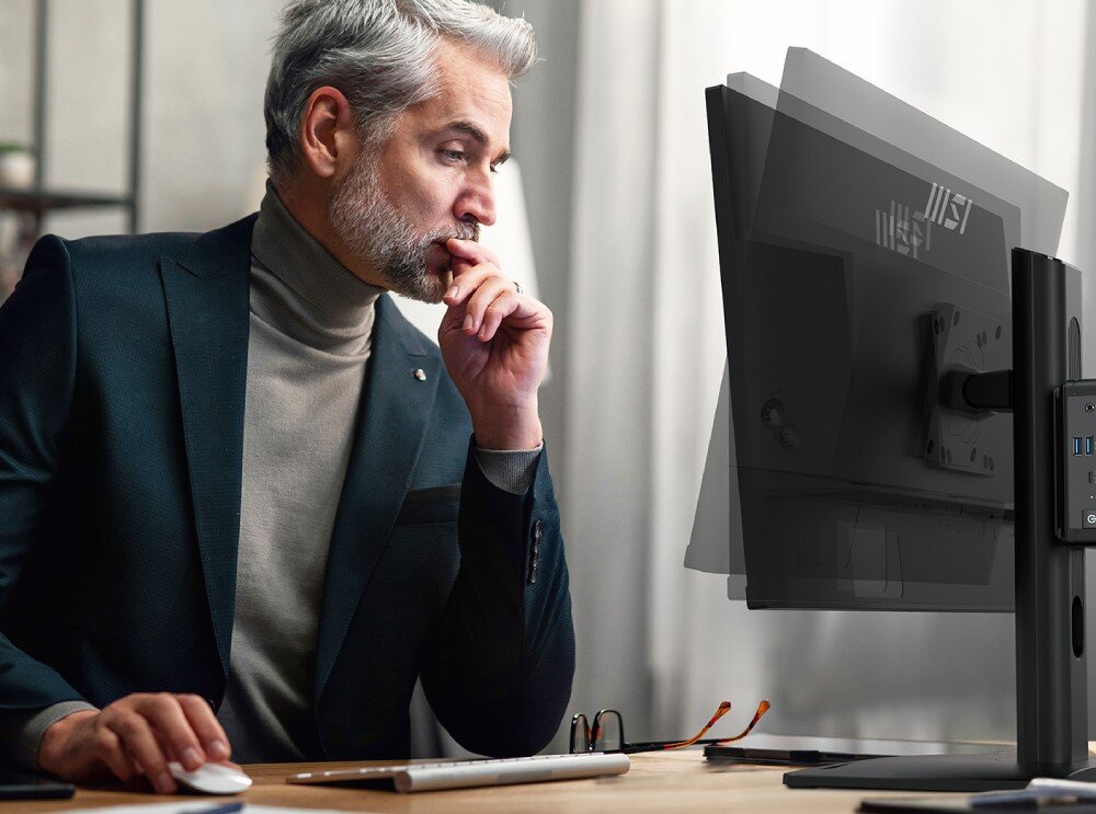 Osoba siedzi przy biurku, patrząc na monitor komputerowy. Monitor jest zamontowany na regulowanym ramieniu, a na jego tylnej części widoczne jest logo MSI.
