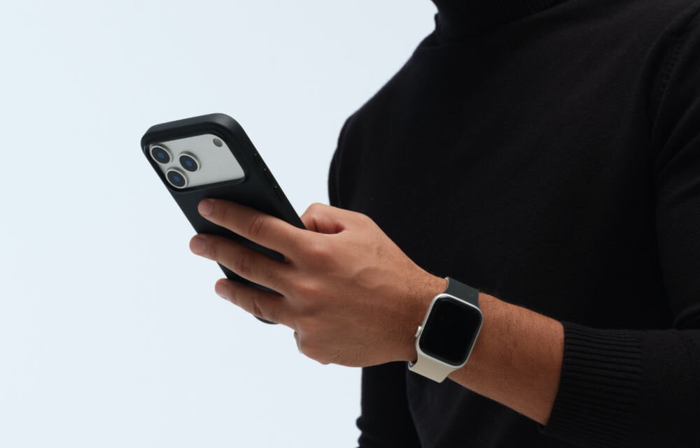 Osoba trzyma smartfon w czarnym etui, na nadgarstku ma zegarek z ciemnym ekranem i jasnym paskiem.