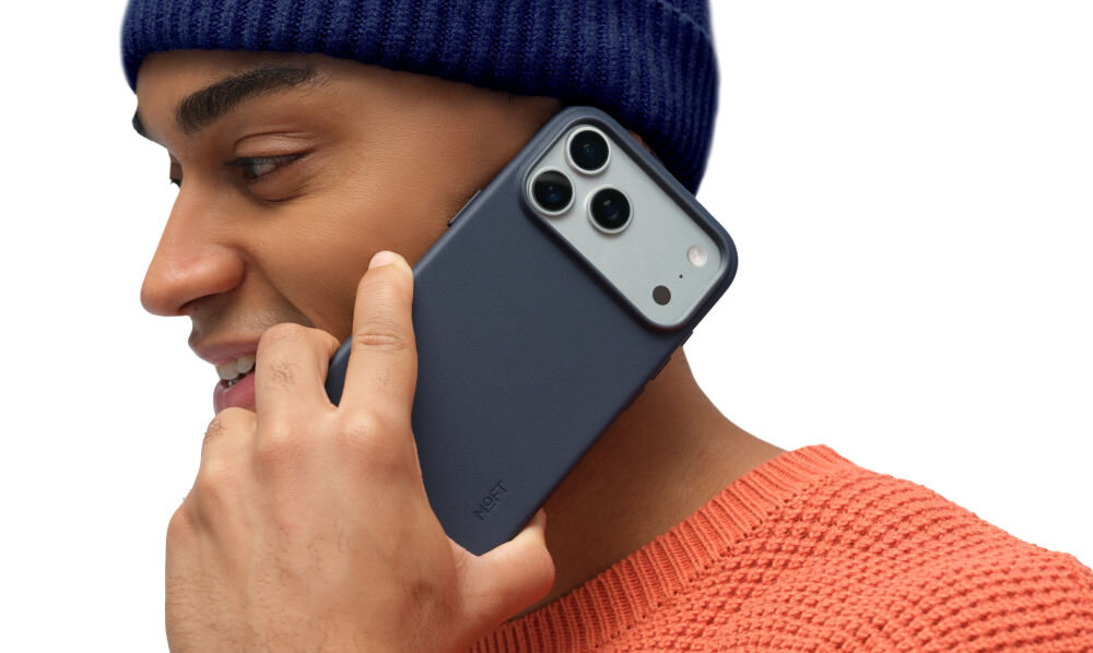 Osoba trzyma przy uchu telefon w granatowym etui z trzema widocznymi obiektywami aparatu. Na etui widoczny jest napis \'MOFT\'.