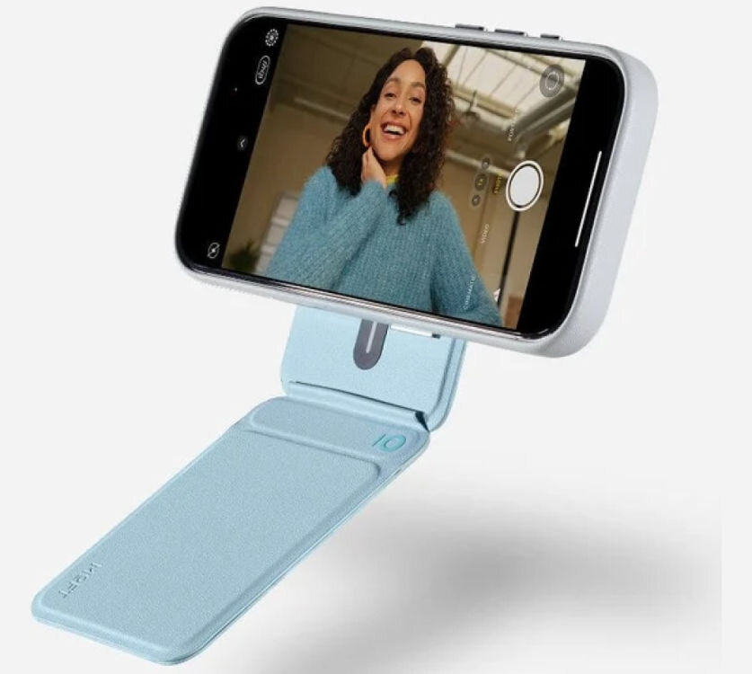 Telefon zamocowany na niebieskim magnetycznym portfelu i stojaku, na ekranie widoczna osoba w niebieskim swetrze. Na portfelu widoczne jest logo \'MOFT\'.
