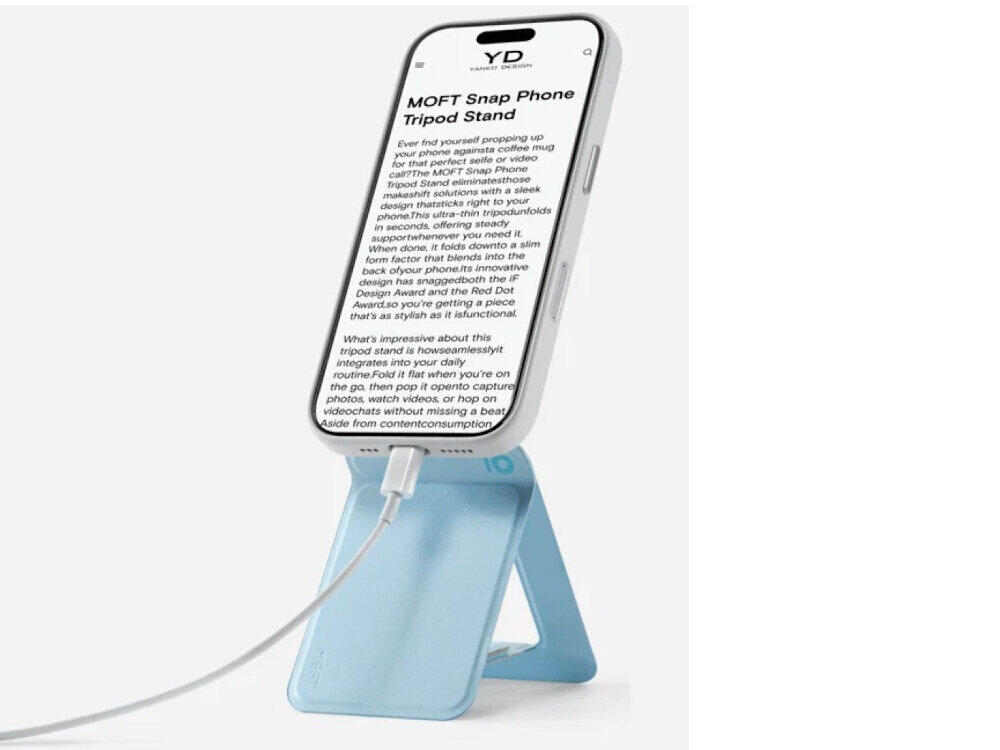 Telefon umieszczony jest na niebieskim stojaku, z podłączonym kablem. Na ekranie telefonu widoczny jest tekst zaczynający się od \'MOFT Snap Phone Tripod Stand\'.