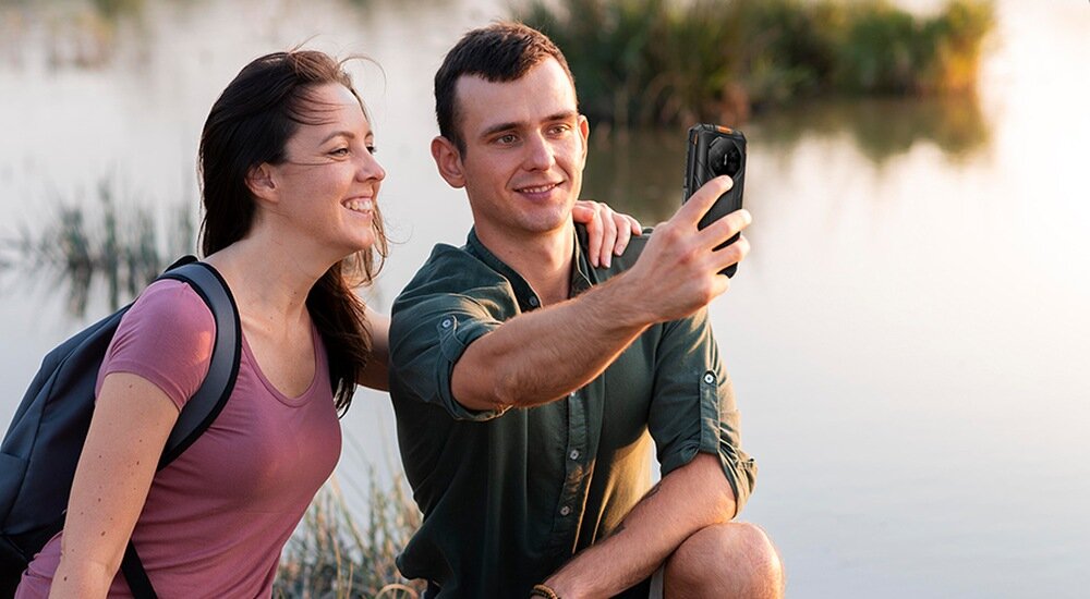 Dwie osoby robią selfie nad wodą, trzymając smartfon. W tle widać roślinność i odbicie w wodzie.