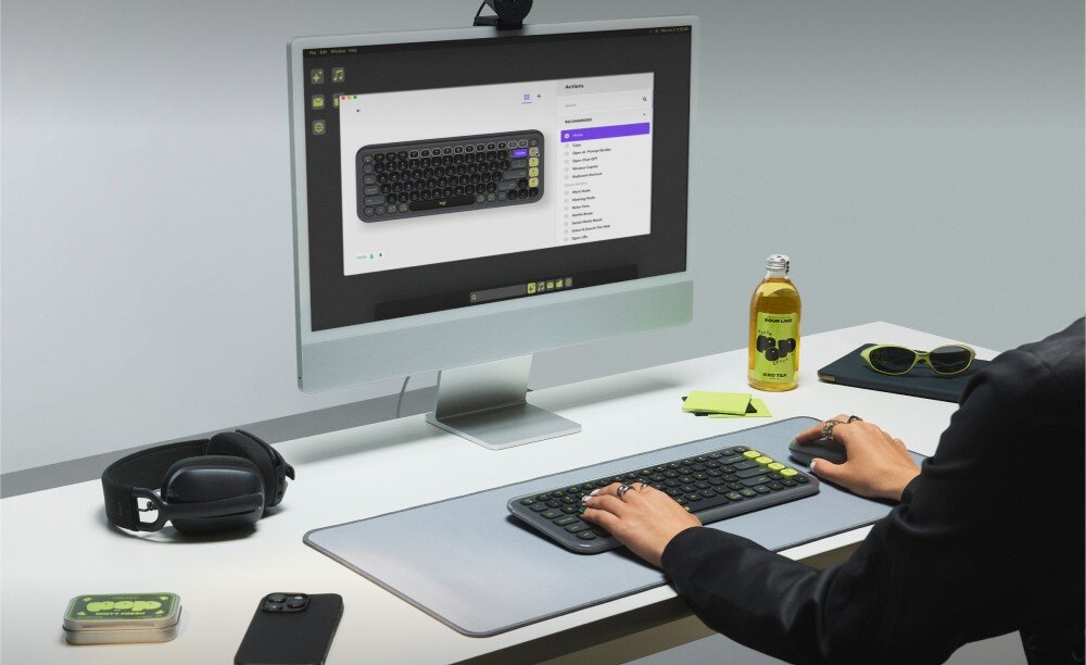 Na biurku znajduje się klawiatura z okrągłymi klawiszami, przed monitorem wyświetlającym jej obraz. Obok leżą słuchawki, telefon, napój i okulary przeciwsłoneczne.