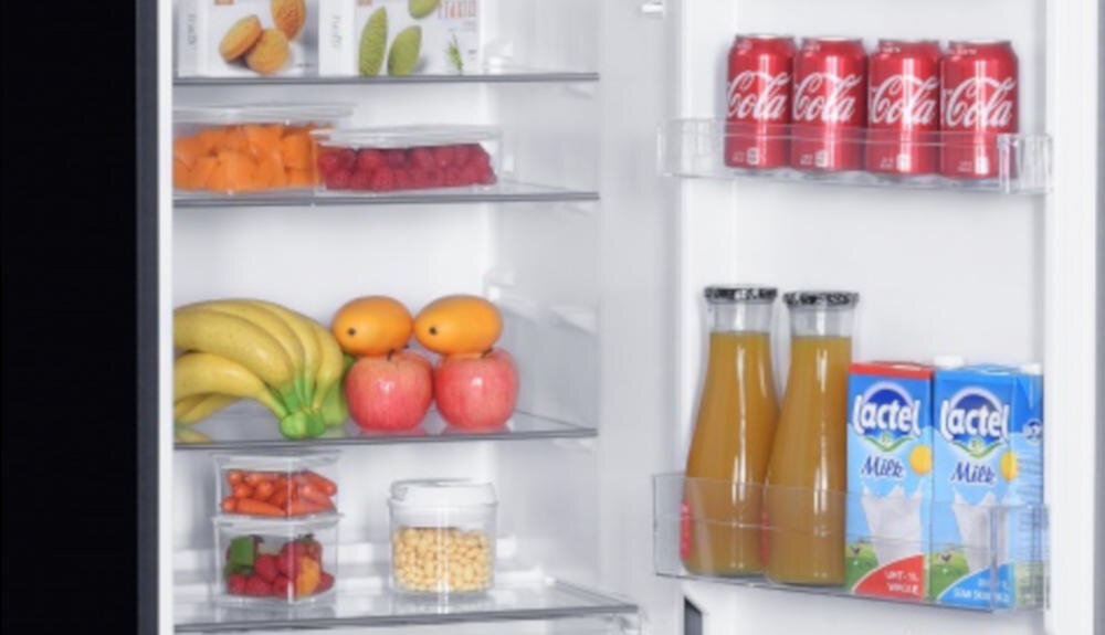 Wnętrze lodówki z różnorodnymi produktami spożywczymi, w tym owoce, napoje i puszki z napojem gazowanym. Na drzwiach widoczne są butelki soku i kartony mleka z logo Lactel.