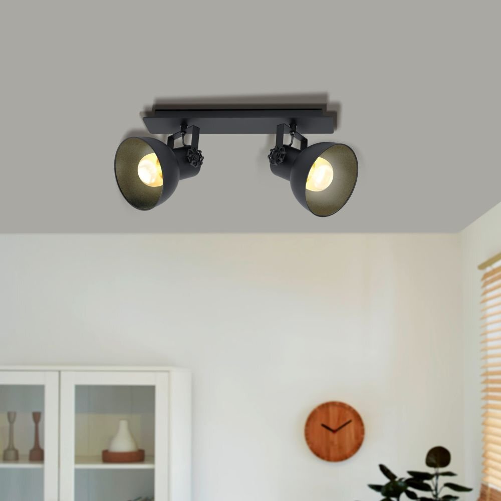 Na suficie zamontowana jest czarna lampa reflektorowa z dwoma źródłami światła. W tle widoczna jest część wnętrza z szafką i zegarem ściennym.