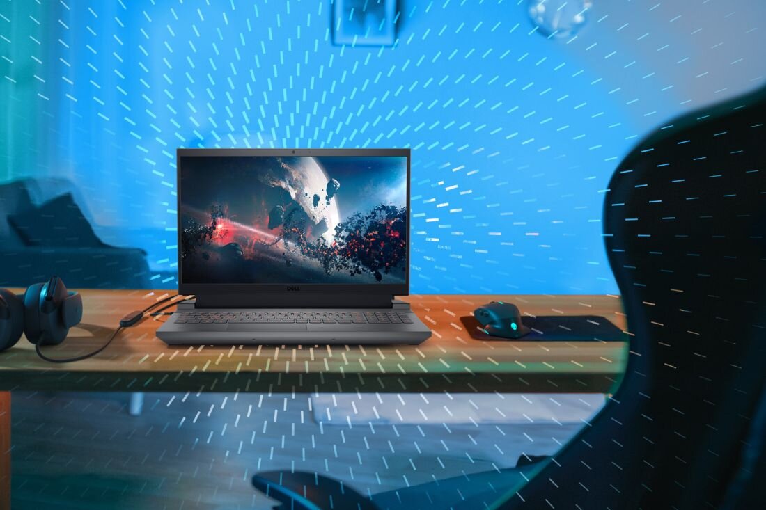 Na biurku znajduje się laptop z wyświetlonym obrazem kosmosu, obok leżą słuchawki i myszka na podkładce. W tle widać rozmyte elementy pokoju i niebieskie linie sugerujące ruch powietrza.