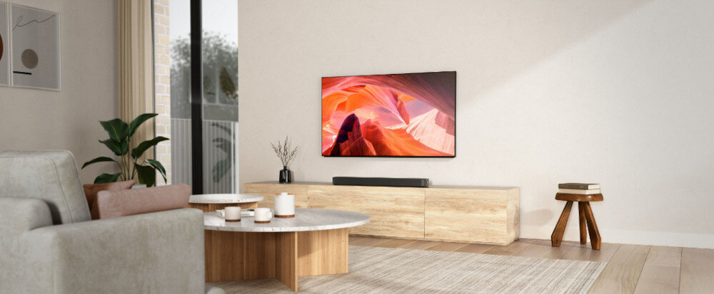 W salonie znajduje się telewizor zamontowany na ścianie, poniżej którego umieszczony jest czarny soundbar na drewnianej szafce. Obok stoi okrągły stolik z kilkoma dekoracjami, a w tle widać roślinę i okno.