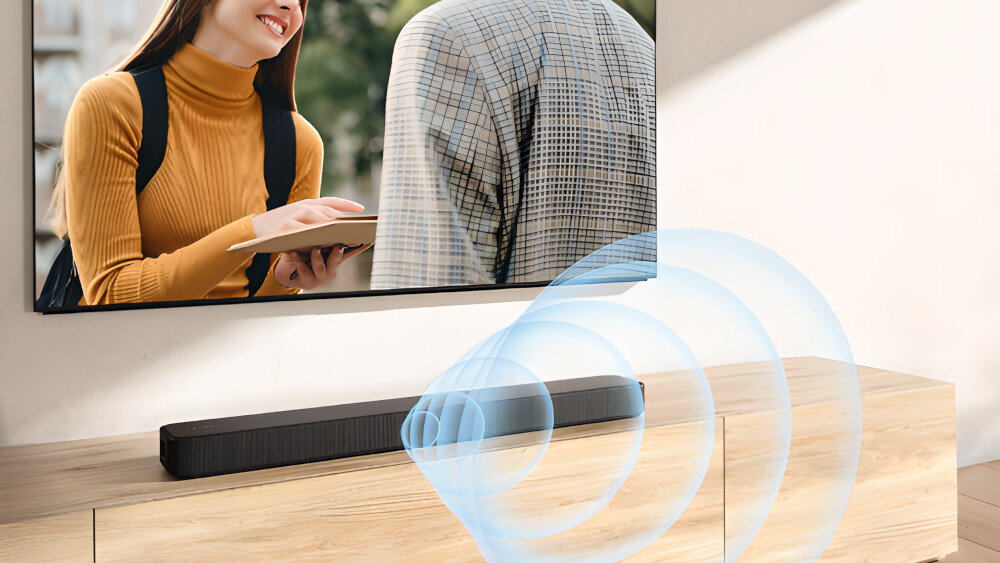 Na drewnianej szafce stoi czarny soundbar, przed którym widoczne są niebieskie fale dźwiękowe. W tle znajduje się telewizor z obrazem przedstawiającym dwie osoby.