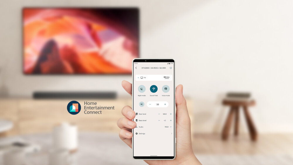 Osoba trzyma smartfon z aplikacją do sterowania dźwiękiem, na ekranie widoczne są opcje regulacji głośności i ustawień. W tle znajduje się telewizor z kolorowym obrazem oraz logo \'Home Entertainment Connect\'.