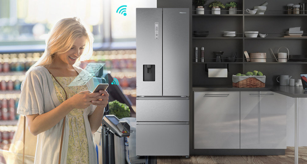 Na obrazie widoczna jest lodówka w kolorze inox z panelem sterowania i dystrybutorem wody na drzwiach. Obok stoi osoba trzymająca smartfon, a w tle widać półki z produktami spożywczymi.