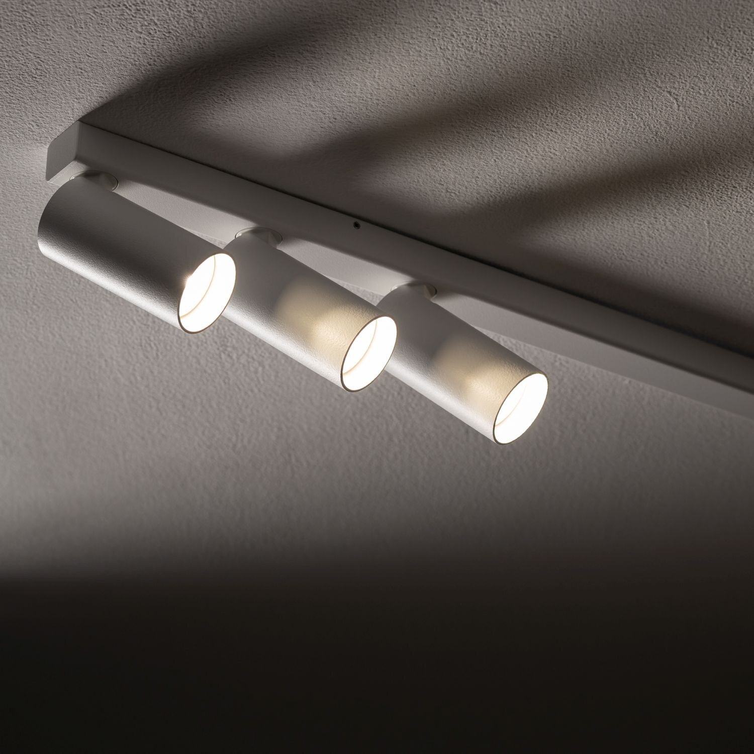 Na obrazie widoczna jest lampa sufitowa z trzema cylindrycznymi reflektorami, które emitują światło. Lampa jest zamontowana na prostokątnej listwie przymocowanej do sufitu.