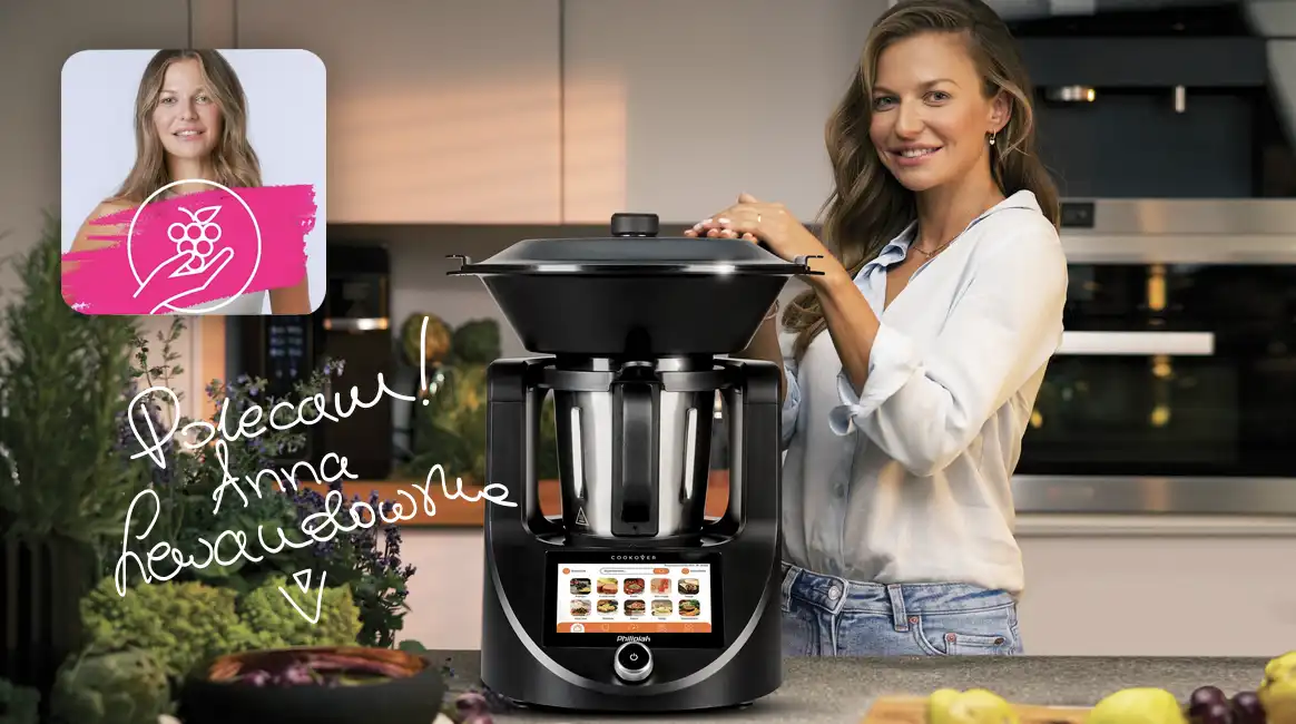 Kobieta stoi obok czarnego robota kuchennego z ekranem dotykowym, na którym widoczne są różne potrawy. Na obrazie znajduje się napis: \'Polecam! Anna Lewandowska\'.