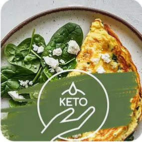 Na talerzu znajduje się omlet z kawałkami sera i liśćmi szpinaku. Na obrazie widoczny jest symbol z napisem \'KETO\'.