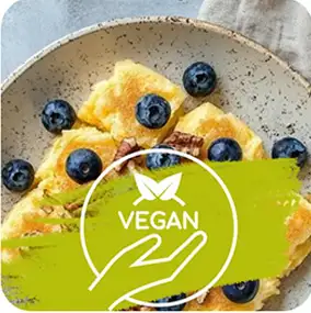 Na talerzu znajdują się kawałki jedzenia z jagodami, a na pierwszym planie widoczny jest zielony symbol z napisem \'VEGAN\'.