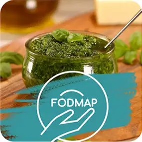 Szklany słoik z zielonym pesto, ozdobiony liśćmi bazylii, stoi na drewnianej desce. Na obrazie widnieje napis \'FODMAP\' z symbolem dłoni.