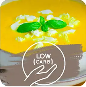 Na obrazie widoczna jest zupa o kremowej konsystencji, ozdobiona listkami bazylii i kawałkami sera. Na dole znajduje się symbol dłoni z napisem \'LOW CARB\'.
