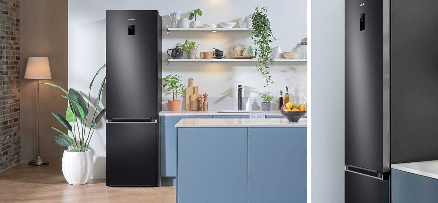 Nowocześnie zaaranżowana kuchnia z lodówką RB38C776CB1, którą możesz kupić w Media Expert Grafitowa lodówka z logo Samsung stoi obok niebieskiej kuchennej wyspy, w otoczeniu roślin i półek z naczyniami. W tle widoczna jest lampa stojąca oraz ceglana ściana.