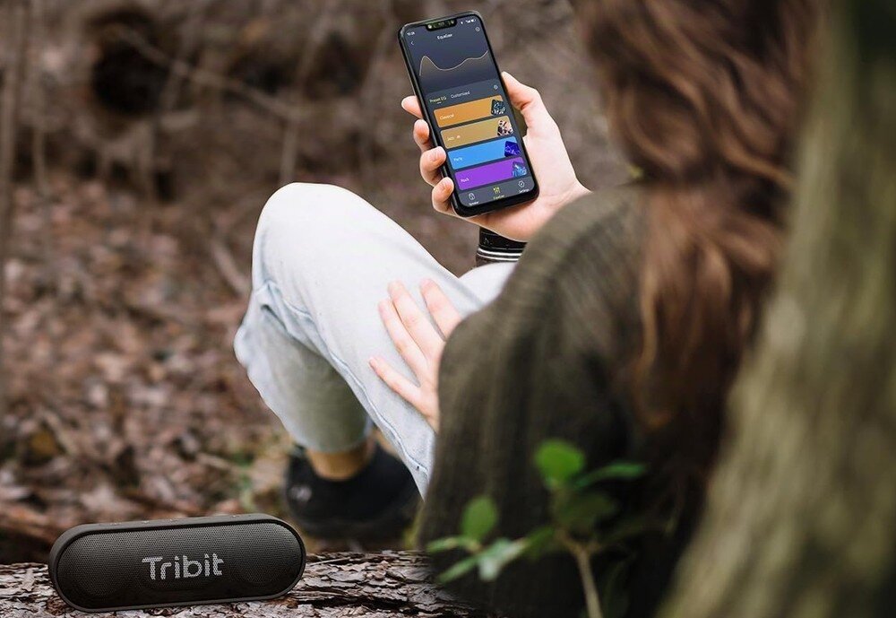 Głośnik mobilny TRIBIT XSound Go BTS58, aplikacja TRIBIT, regulacja dźwięku, mikrofon, asystent głosowy, osoba trzymająca telefon w ręku , głośnik stojący na pniu