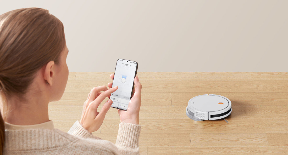 Na drewnianej podłodze znajduje się biały robot sprzątający. Osoba trzyma smartfon z aplikacją do sterowania robotem.